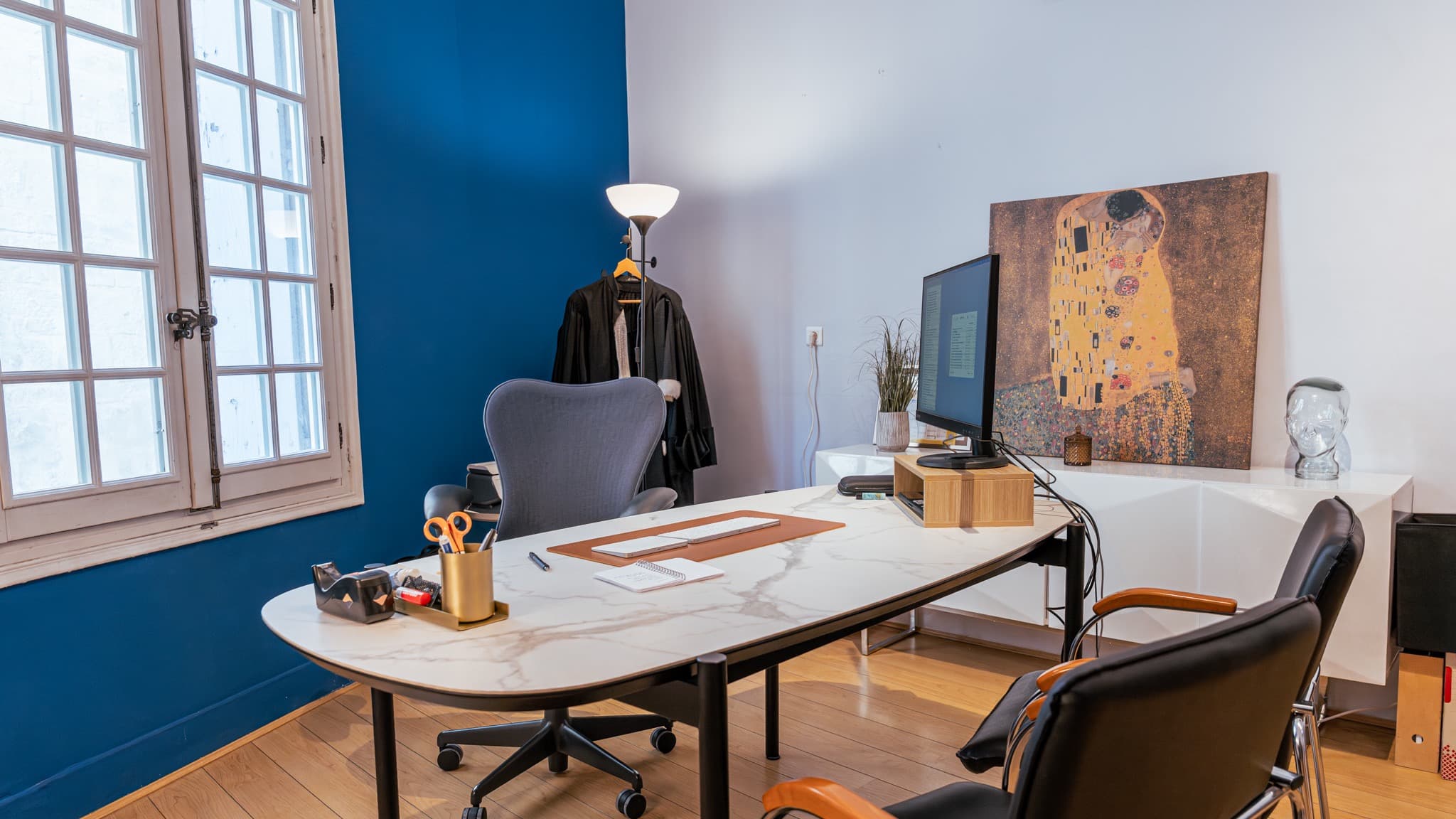 Bureau bleu profond dans un immeuble ancien de l’Écusson, avec une robe d’avocat suspendue près d’une fenêtre à petits carreaux à ferronnerie d’époque, un bureau ovale à plateau de marbre blanc et un parquet en chêne clair.